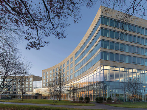 The curved modern architecture of Tata Hall at Harvard Business School showcases multiple floors with expansive glass windows reflecting the clear blue sky. Bare trees and a green lawn surround the building, captured at dusk when interior lights begin to glow.