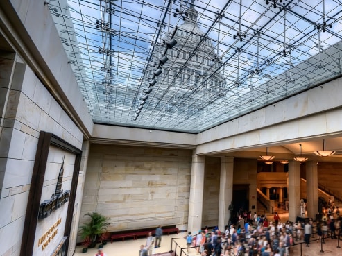 The spacious atrium at the Eaton corporate headquarters features a glass ceiling revealing the dome of the U.S. Capitol. People gather in the foreground, with a staircase and palm plant on the left. Marble surfaces enhance the elegant atmosphere.