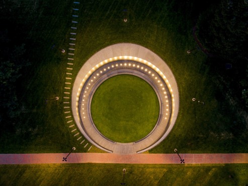 Aerial view of a circular structure crafted from Vals Quartzite with a ring of lights, surrounded by lush green grass. A red brick pathway crosses in front, and the scene is softly lit, creating a serene atmosphere at dusk.