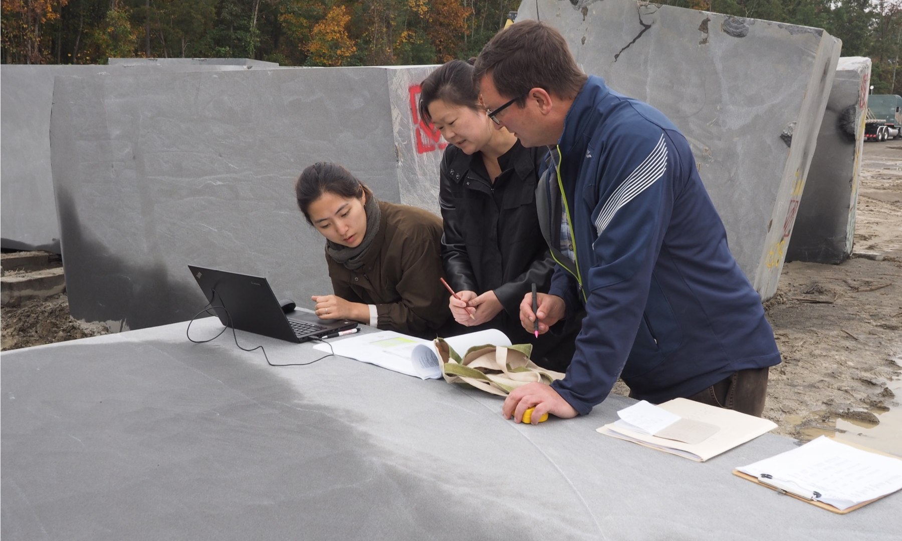 Three people stand outdoors examining documents and a laptop atop large stone slabs at the Collier Memorial. Two women and a man are engaged in discussion, surrounded by industrial blocks, focused on what seems to be a construction or architectural task.