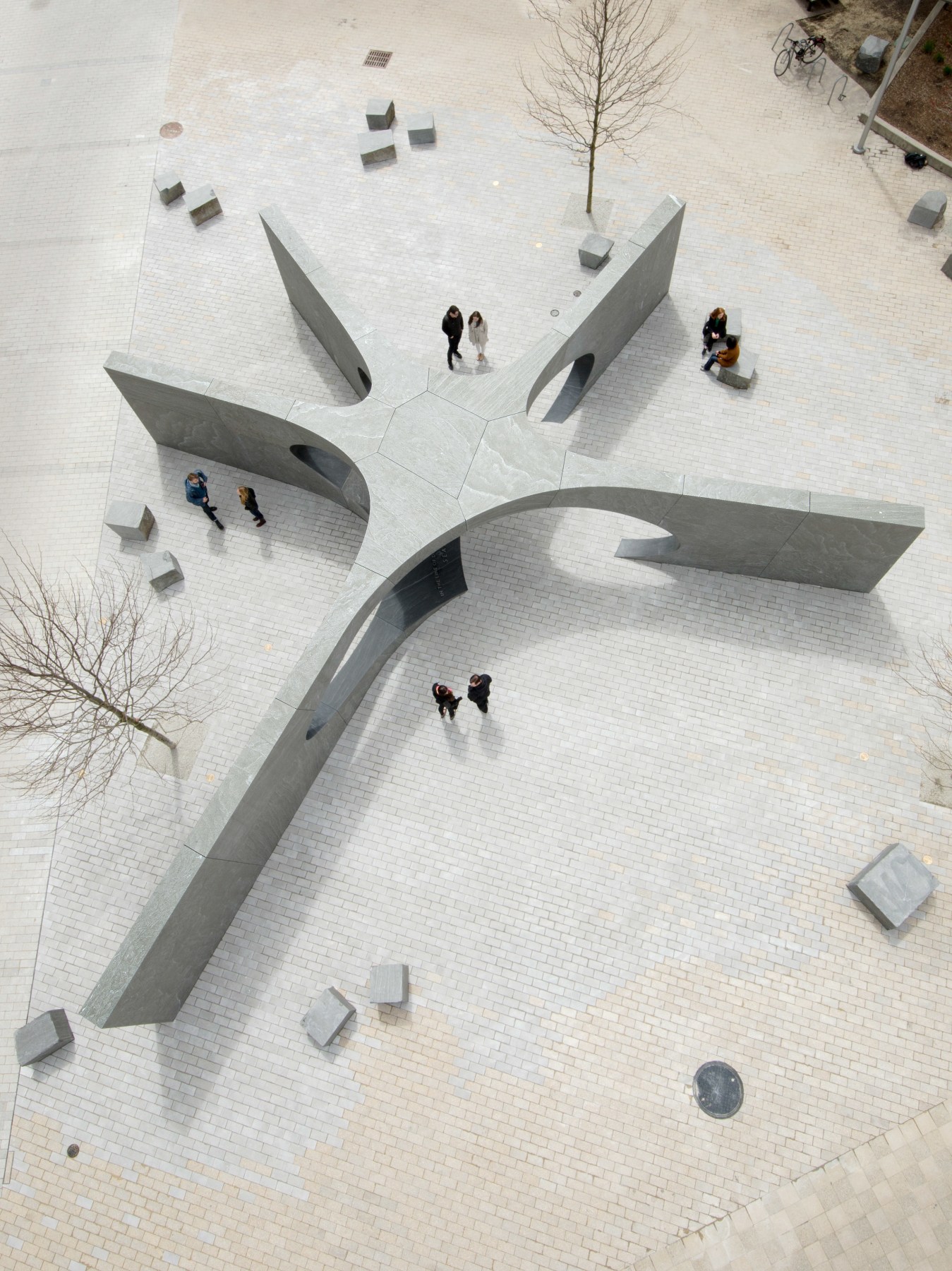 An aerial view reveals the Collier Memorial, a modern, star-shaped concrete sculpture in a plaza. Intersecting arches and walls create its striking design as people stroll and gather around on the light-colored tiles. Leafless trees and scattered square benches complete the scene.