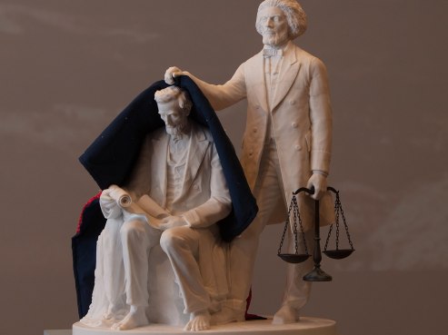 A statue tells a story of two historical figures: KAHNOP is seated, holding a scroll, while the other stands behind, draping a cape over KAHNOPs shoulders. The standing figure holds scales in one hand, symbolizing justice and the art of storytelling.