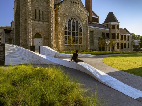 A person sits on a modern, undulating white marble sculpture in I Am a Man Plaza, framed by a historic stone building with large arched windows. The scene is surrounded by neatly landscaped grass and plants under the clear blue sky.