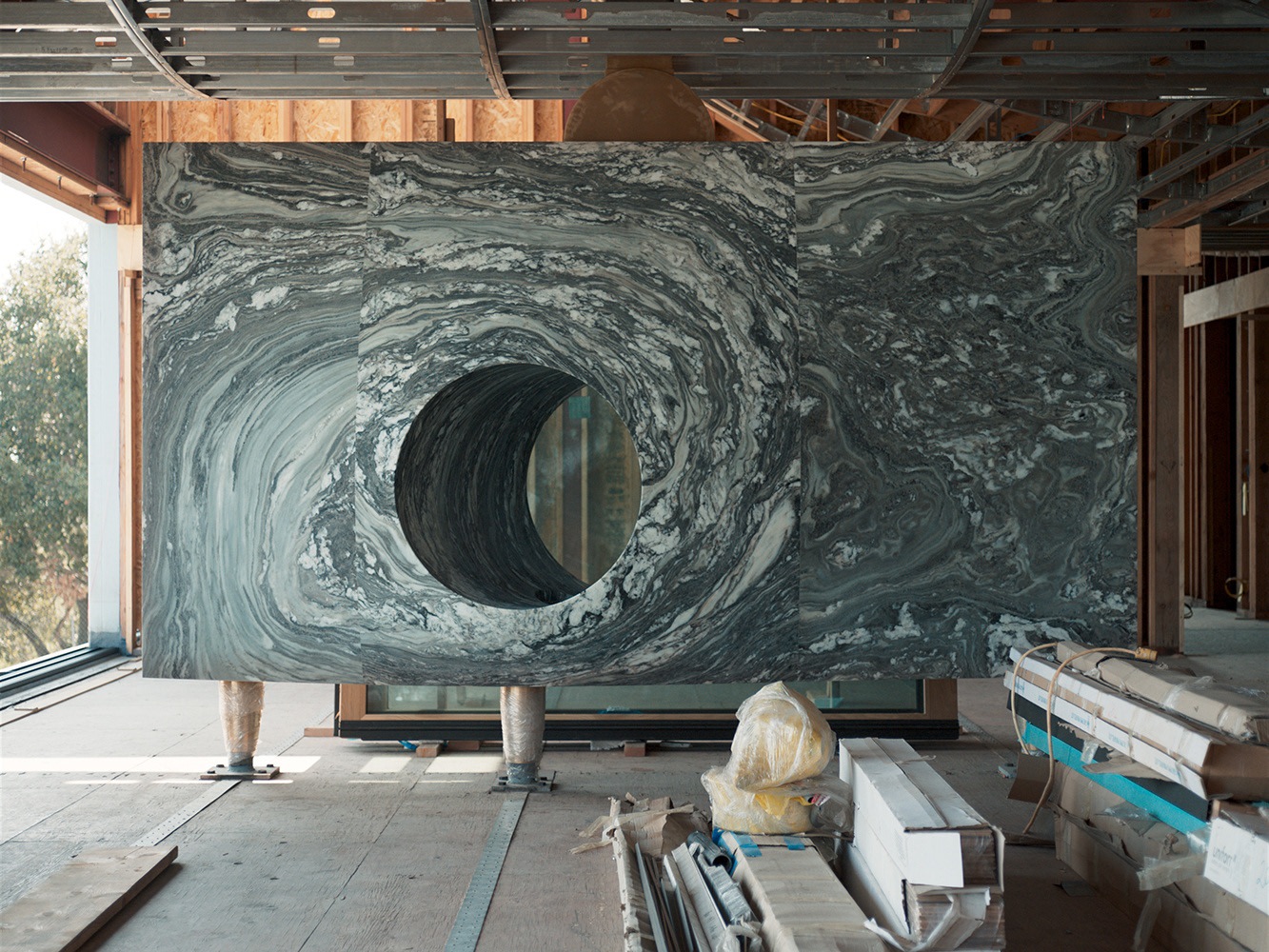 A large, swirling gray and white marble slab with a circular cutout stands in the partially constructed House of Horns by WOJR. Construction materials and wooden beams surround the area, while natural light streams in through an open section.