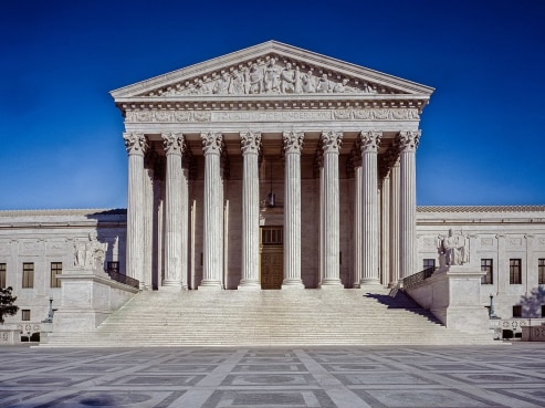 The image captures the majestic United States Supreme Court building in Washington, D.C. Its neoclassical facade showcases a row of tall columns beneath a grand pediment. Stairs ascend to the entrance, all set against a backdrop of clear blue sky.