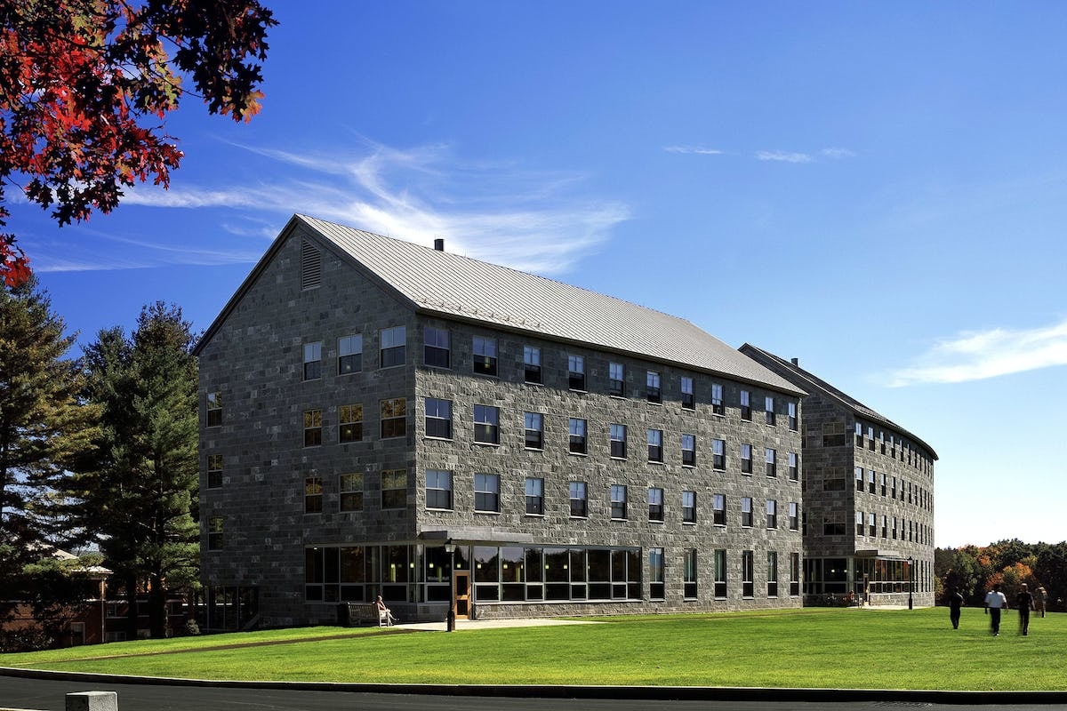 A large stone building with several windows stands on Amherst Colleges grassy lawn under a clear blue sky. Trees with vibrant autumn foliage surround the structure, where people are walking nearby.