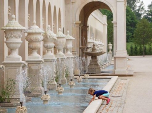 A young child, visiting the Eaton corporate headquarters, leans over a long, decorative fountain lined with ornate urns and jets of water. The stone colonnade arches above, while lush greenery peeks out in the background. The child sports a blue shirt and red shoes.