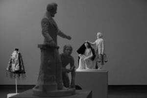 Black and white photo of a museum exhibit, titled Lifting the Veil, featuring sculptures: a standing figure, a kneeling person, and a partially draped seated figure holding scales. Nearby, a decorated jacket adds contrast to this evocative scene.