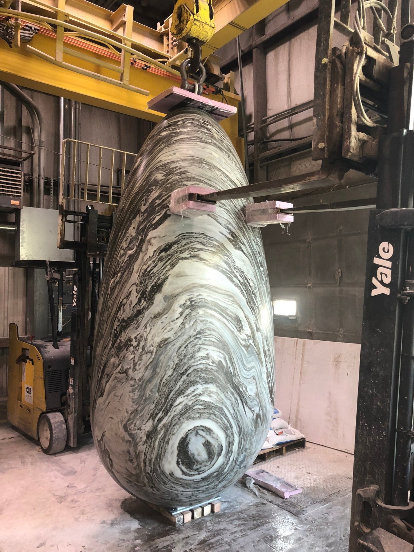 Inside an industrial building, a large, oval-shaped sculpture adorned with swirling gray and white patterns is carefully lifted by a crane. This masterpiece, known as the House of Horns by WOJR, is surrounded by various pieces of equipment and machinery.