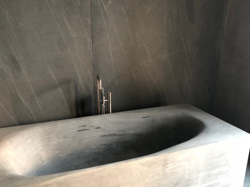 A sleek, stone bathtub with an integrated faucet stands against a dark, textured wall in the heart of a 28th Street Penthouse. The minimalist design and neutral tones create a modern, luxurious bathroom ambiance that epitomizes sophisticated NYC living.