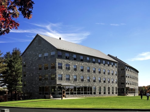 A four-story stone building with a peaked roof and penthouse, featuring numerous windows. It’s set against a blue sky with scattered clouds. A grassy lawn and trees frame the scene, with a few people strolling nearby.