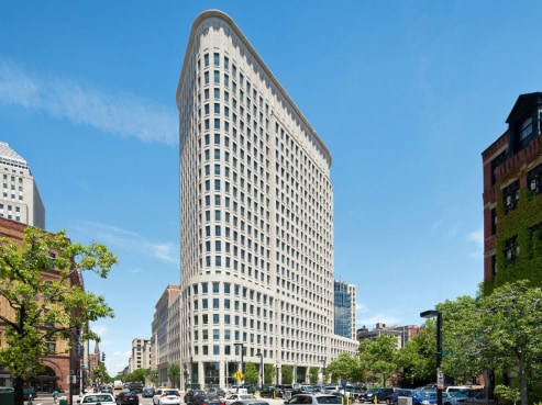 A tall, curved skyscraper, serving as the headquarters for Liberty Mutual Insurance, stands under a clear blue sky. It is surrounded by smaller buildings and trees, with cars bustling on the street below.