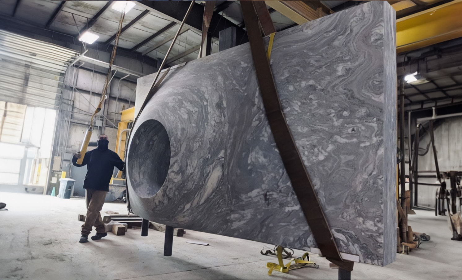 In a warehouse reminiscent of Pietrasantas artistry, a worker deftly guides a large slab of gray marbled marble with a circular cutout. The slab, suspended by straps attached to a crane, hangs beneath the metal roofing and fluorescent lights, evoking the craftsmanship of Italy.