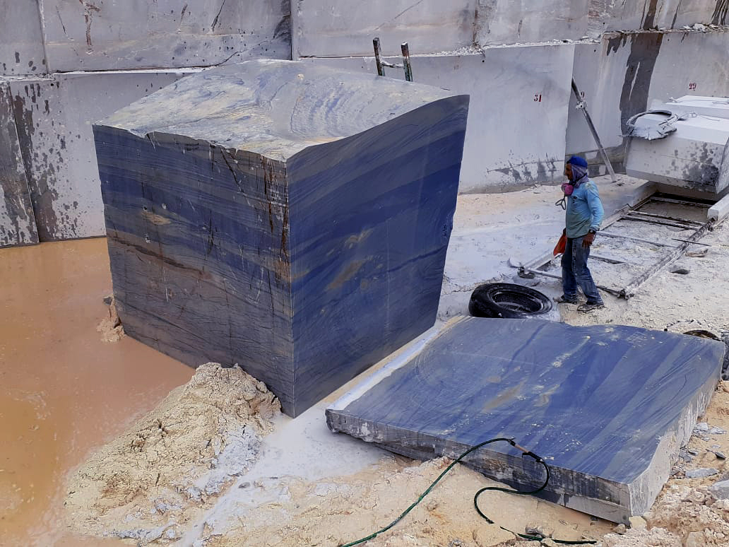 A person stands near a large, cube-shaped block of blue stone and a separated slab in a quarry, surrounded by muddy water, stone walls, and quarry equipment—a snapshot of professional Stone Sourcing in action.