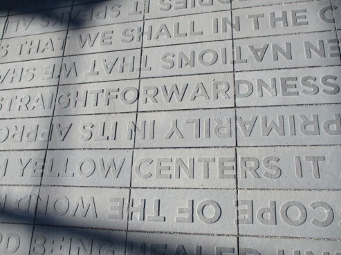 Close-up of a stone pavement with large, engraved letters arranged in lines, telling an upside-down story with partial phrases visible. Shadows cast across the surface, adding contrast to the textured gray background and whispering tales of KAHNOP.