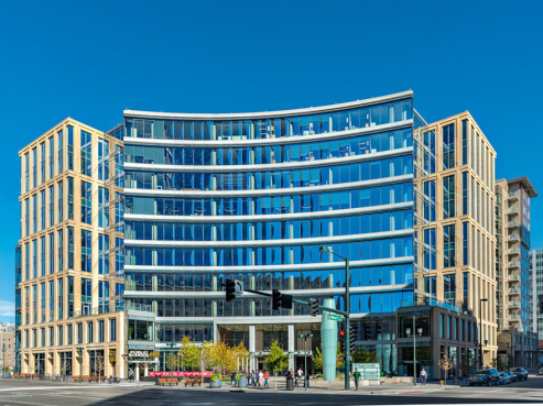 Located at 1601 Wewatta Street, this modern multi-story office building boasts a curved glass facade that mirrors the clear blue sky. Tan vertical columns add contrast, while a nearby street intersection features traffic lights and a handful of pedestrians in motion.