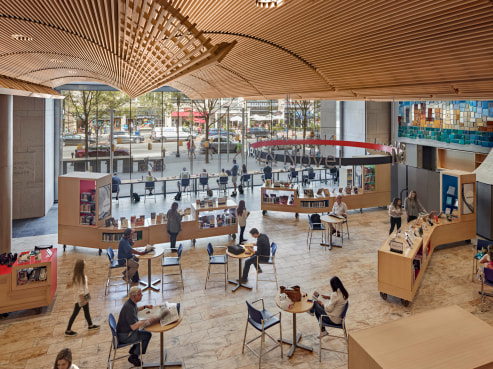 A modern library interior with high wooden ceilings and large windows evokes the elegance of Boston Public Library. People are seated at round tables reading and working, surrounded by shelves of books and magazines. Outside, trees line a bustling street filled with pedestrians.