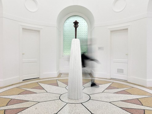 A blurred person walks past a tall, white cone-shaped sculpture in a room with KAHNOPs signature star-like floor pattern. The neutral tones tell a story of elegance against the backdrop of white walls and two doors. In the background, a green, patterned window adds a splash of intrigue.