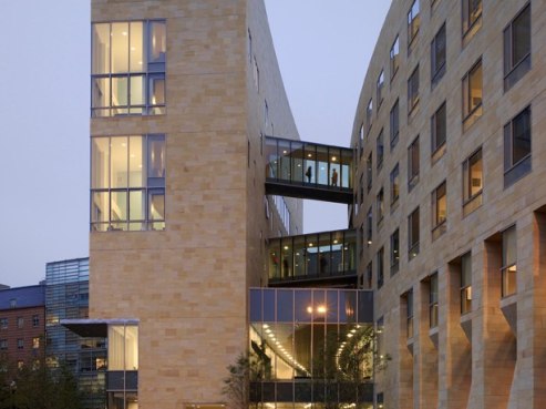 Located at 1601 Wewatta Street, this modern building boasts a beige stone facade with expansive glass windows. Two sleek glass pedestrian bridges connect the main structure to a smaller adjacent one. Interior lights glow warmly against the overcast evening sky.