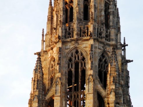 A tall, ornate Gothic bell tower with intricate stonework and pointed arches stands majestically, bathed in warm sunlight against a clear sky. Echoing the grandeur of the Boston Public Library, it boasts elaborate architectural details and decorative spires.