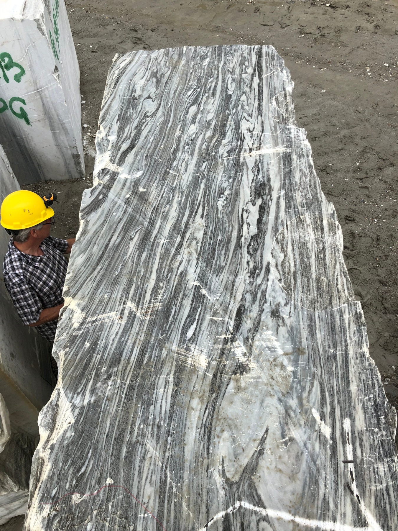 A large, rectangular slab of marble with gray and white swirling patterns is positioned vertically. In the stone yard, a person in a yellow hard hat and plaid shirt stands partially visible beside what could be a future centerpiece for WOJRs intriguing House of Horns project.