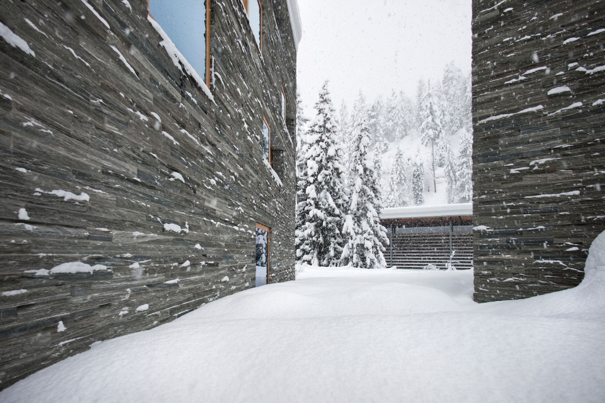 Snow-covered landscape between two stone buildings with wooden windows. Snow blankets the ground and rooftops. In the background, snow-laden pine trees are visible against a snow-filled sky.