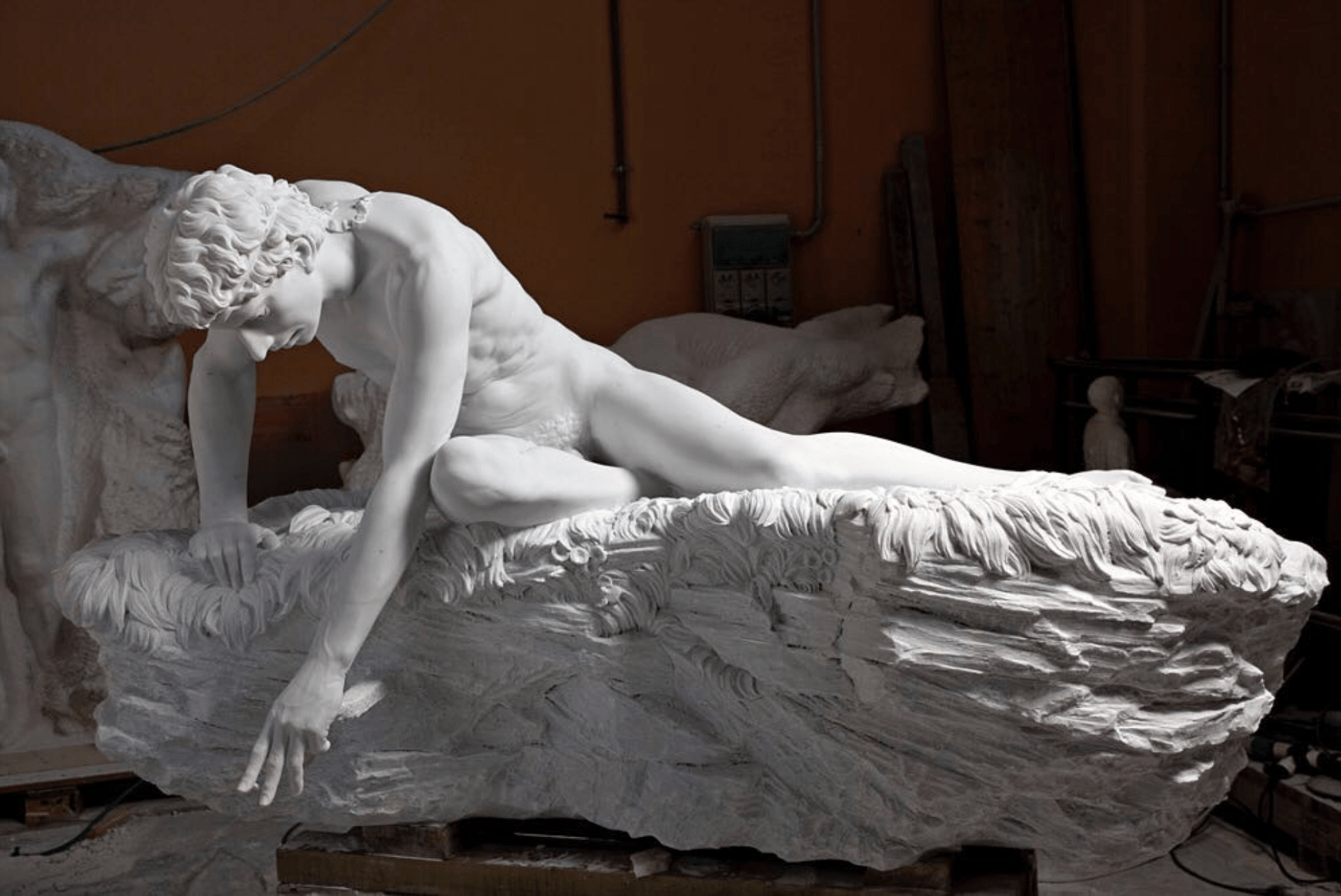 A marble sculpture from Pietrasanta, Italy, portrays a reclining male figure with flowing hair gracefully draped over a textured rock. The detailed piece is set in a workshop, surrounded by tools and equipment.