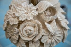 Close-up of an intricate stone sculpture at Longwood Gardens, featuring various flowers such as roses and lilies. The detailed petals and textures reflect the artistry found in horticulture. A slightly blurred blue surface enhances the captivating scene.