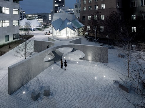 At dusk, the I Am a Man Plaza showcases modern geometric concrete structures with circular cutouts. Two people stand on the paved ground, surrounded by buildings illuminated by urban lighting, highlighting the plazas contemporary design.