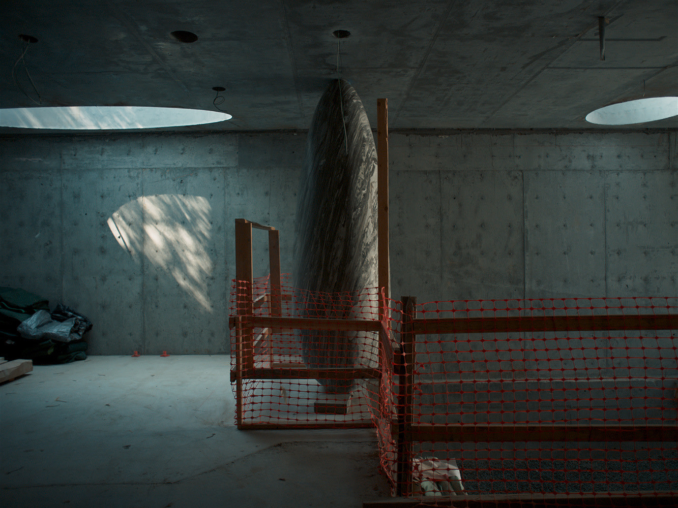 A dimly lit room with concrete walls and ceiling hosts WOJRs enigmatic House of Horns, a large, dark, boat-shaped object standing upright. Enclosed by a wooden frame and orange safety netting, circular skylights cast soft light on the scene. Construction materials lie nearby.