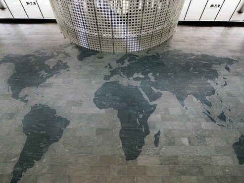 A large, detailed world map is etched on a tiled floor beneath Eatons modern, cylindrical corporate headquarters featuring a sleek grid pattern. The maps dark shades contrast with the light tiles as bright natural light cascades from above.