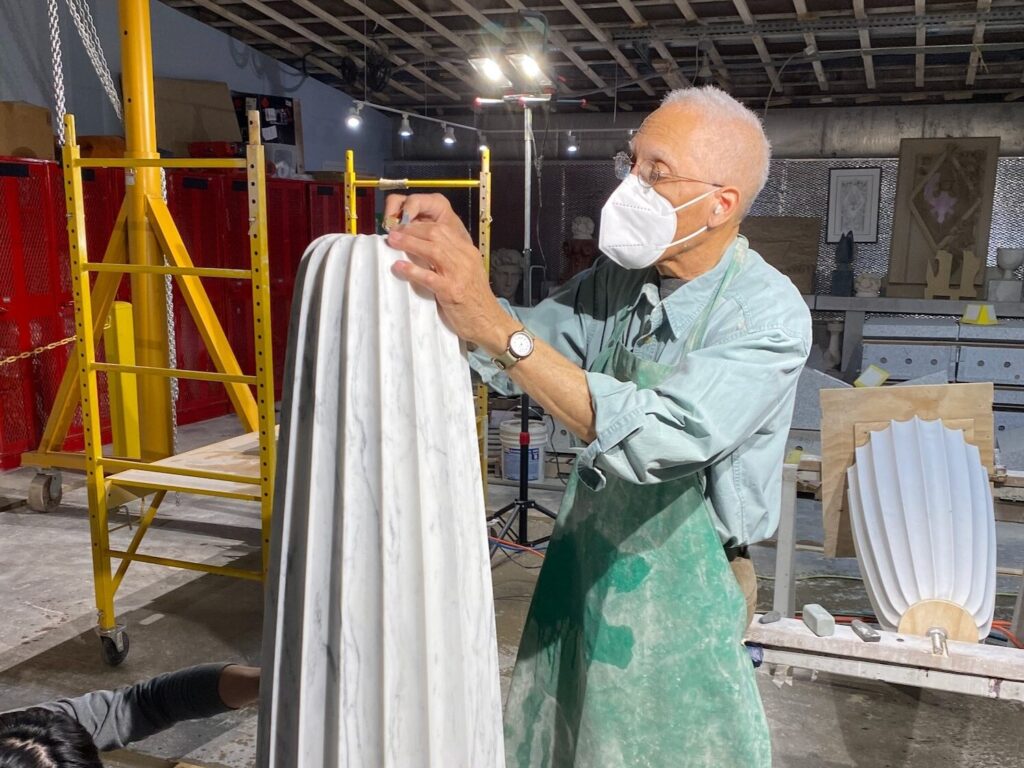 An elderly man, wearing a face mask and apron, carves a tall, textured sculpture resembling a historical figure in his workshop. Surrounded by tools, a yellow ladder, and red lockers in the well-lit industrial space, he crafts with precision and passion.