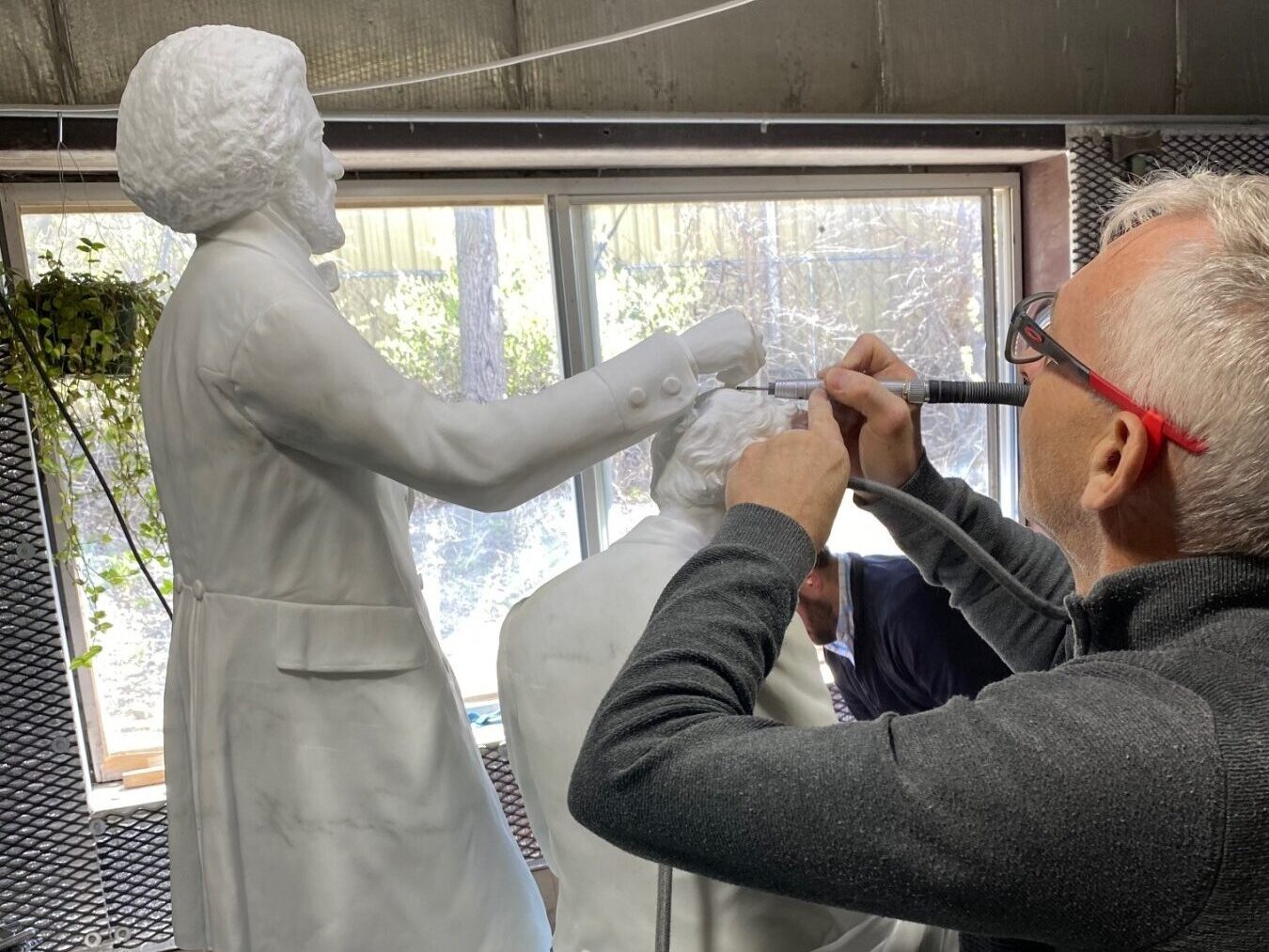 An artist, lifting the veil of creativity, works meticulously on a large marble sculpture depicting two figures. The well-lit studio bathes in natural light from the window. Wearing glasses and dressed in dark clothing, the artist focuses intently on the intricate carving.