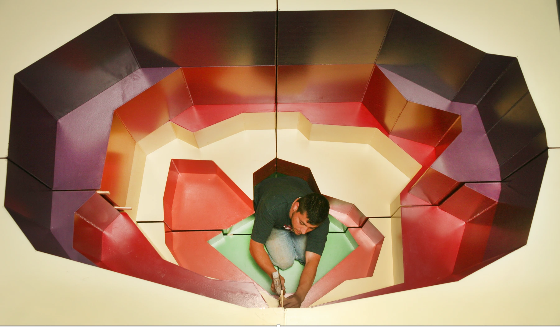 A person is kneeling inside a large, geometric, multi-colored 3D structure resembling an abstract flower. The structure has varying shades of red, purple, and orange. The person appears to be working or assembling the piece, viewed from above.