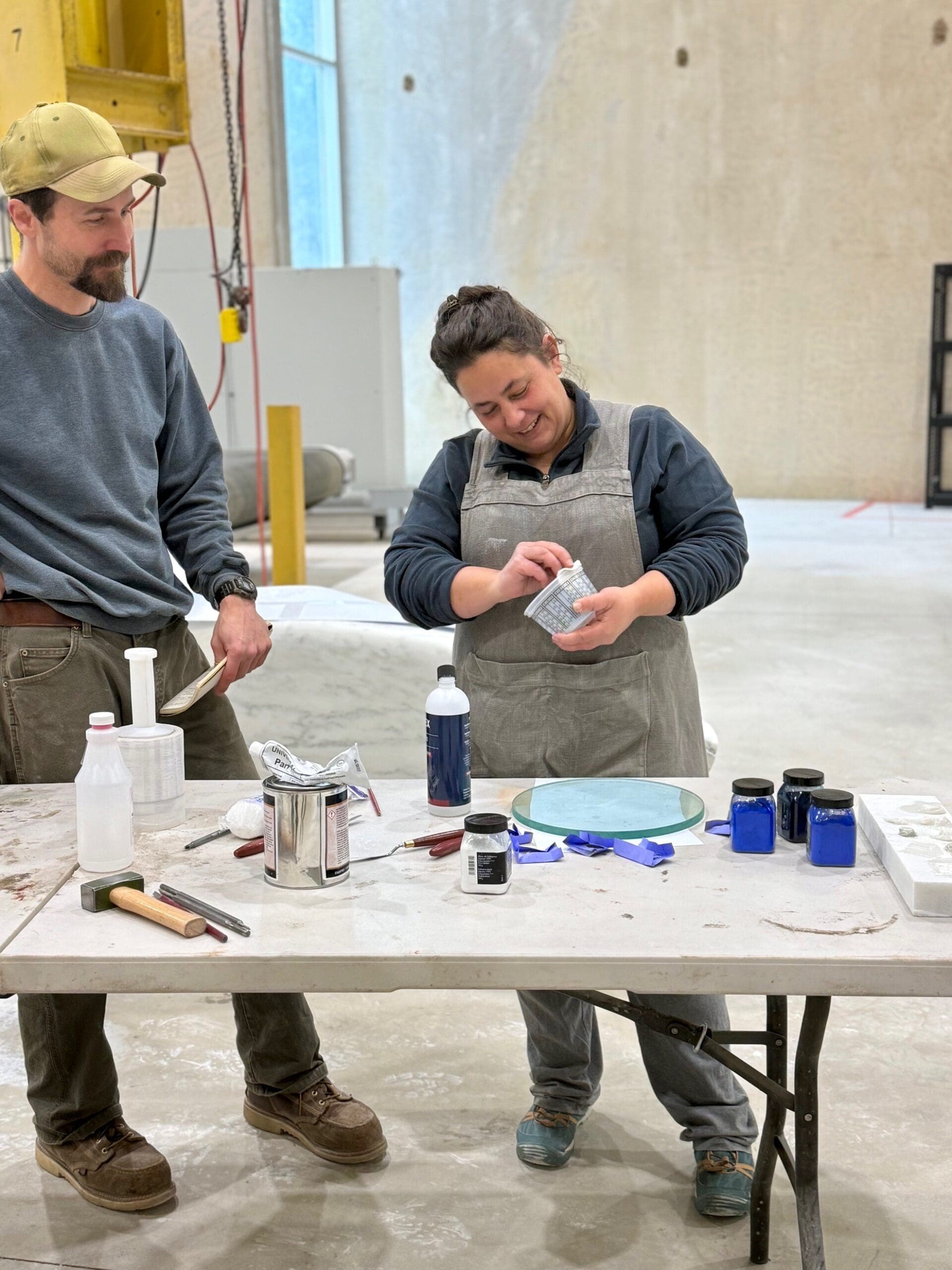 In a bright studio in Pietrasanta, two people stand at a table with art supplies; one woman in an apron mixes something in a small jar while a man in a cap looks on, surrounded by bottles, containers, and blue pigments. Mentorship Spotlight: Rachele Pierini.