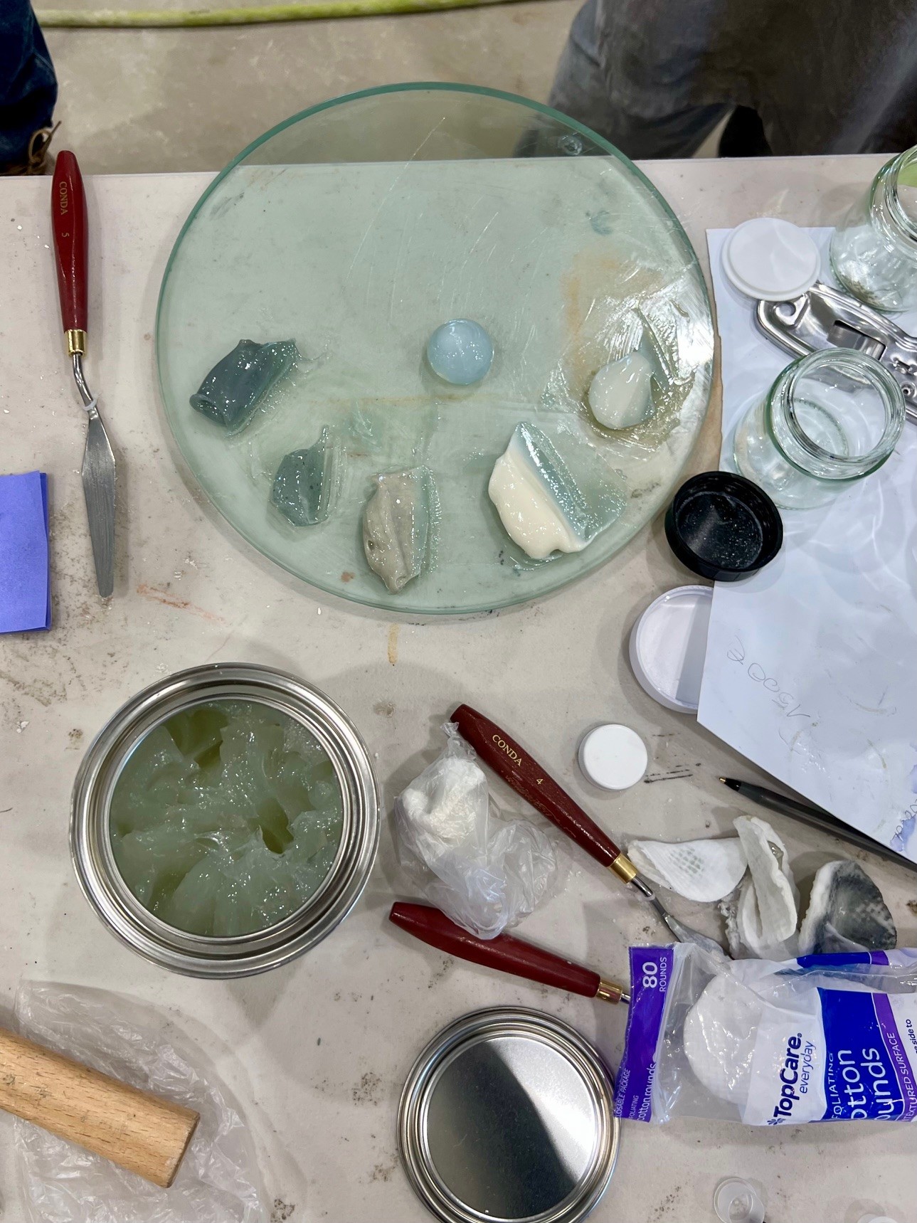 Top-down view of a worktable in Pietrasanta with paint knives, a glass palette holding blobs of pigment, an open can with translucent substance, jars, a mallet, paper towels, and art supplies scattered—perfect for creative mentorship.