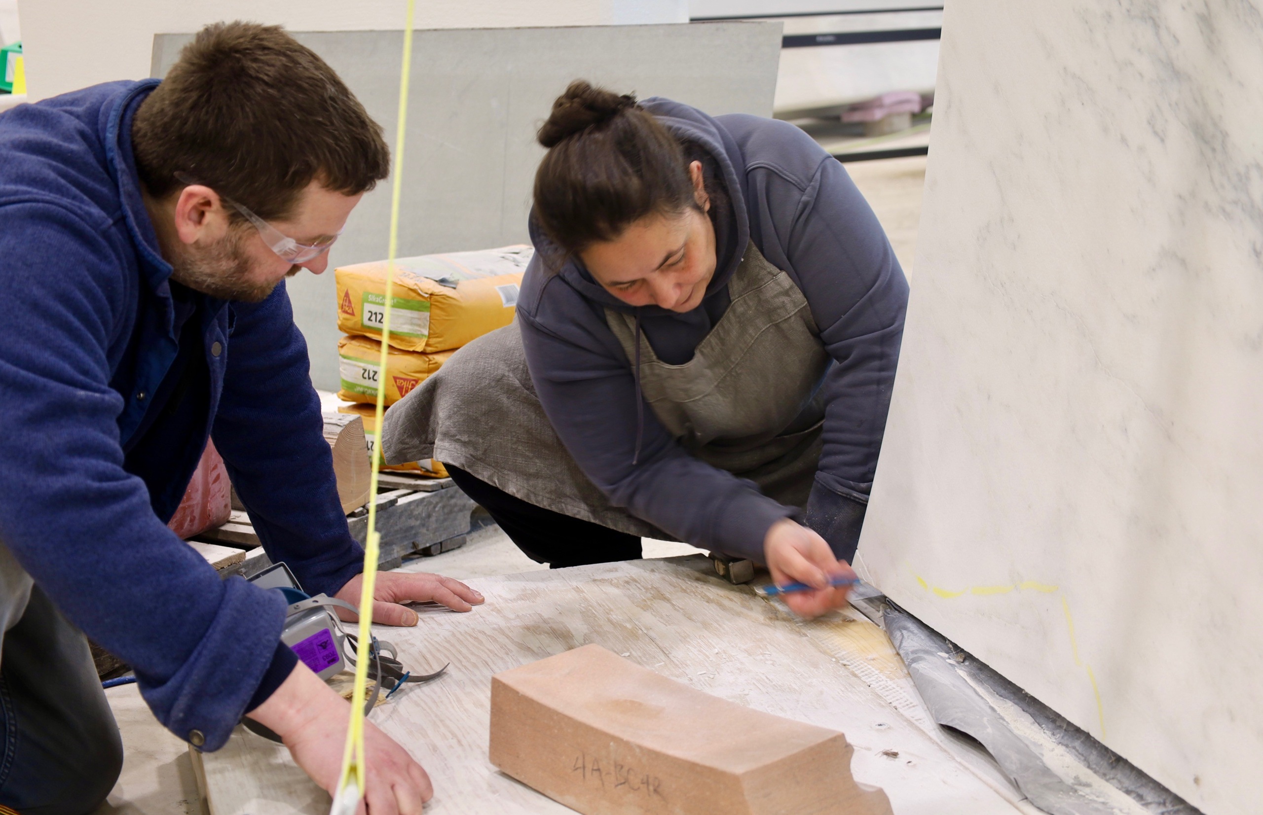 In a Pietrasanta workshop, two people in work clothes measure and mark lines on a large white stone slab; one kneels while the other leans over, both focused on their task amid tools and materials—a true Mentorship Spotlight.