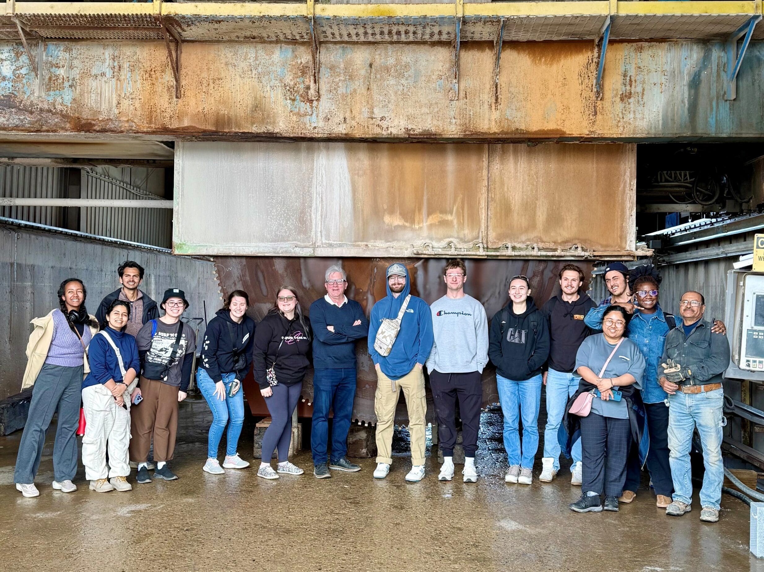 A diverse group of 15 people stands smiling in front of a large industrial structure, part of the Sustainable Design Project. They are casually dressed, with several wearing jackets and sneakers. The background features rusted metal and machinery from the Iowa State Architecture initiative. The ground is wet.