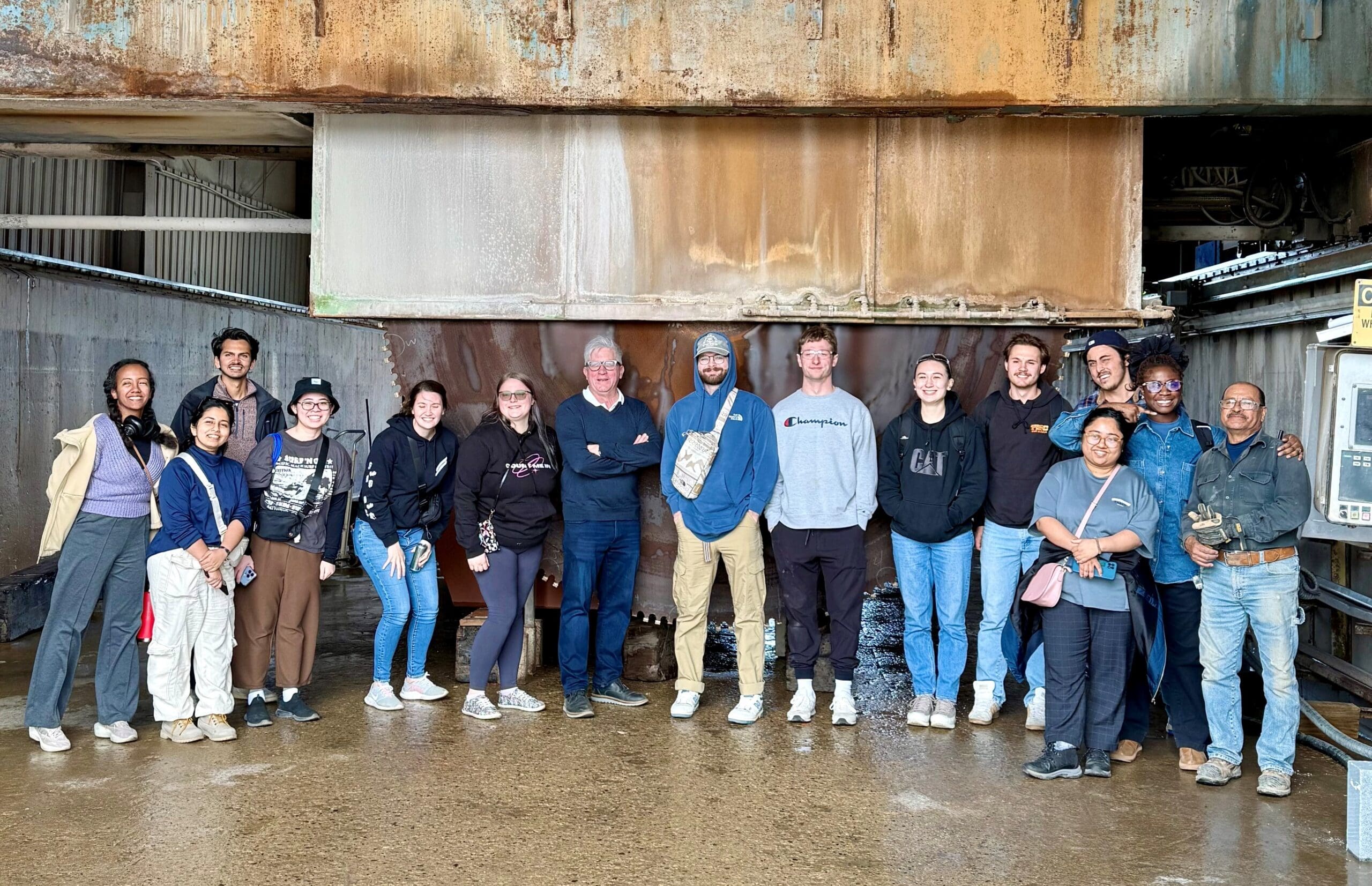 A diverse group of fifteen people stand side by side in an industrial setting with rusty metal walls, showcasing elements of sustainable design. They pose for a photo, most smiling, with the wet ground reflecting their shared vision. Some have their hands in their pockets.