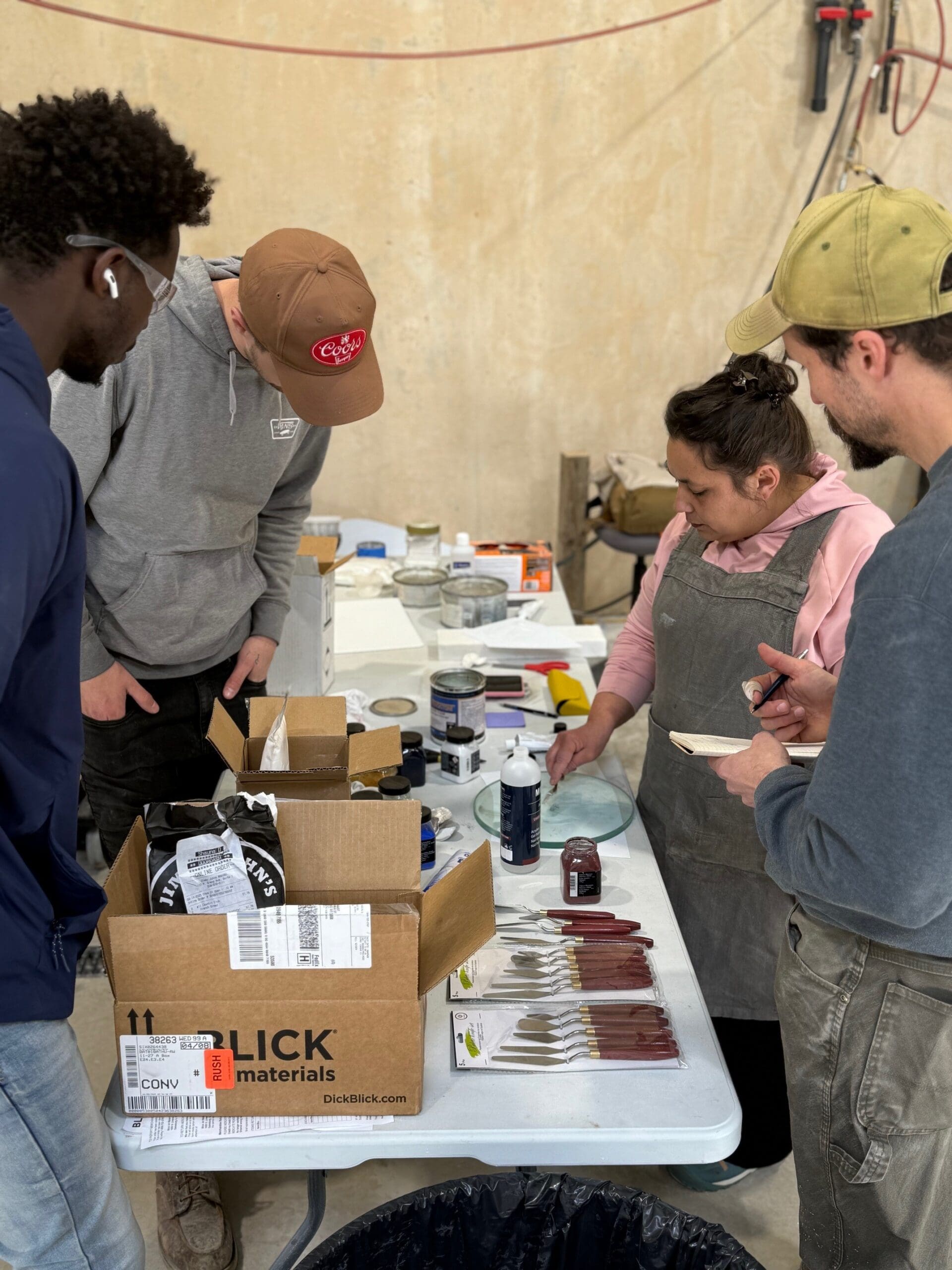 Four people gather around a table with paintbrushes, jars, and art supplies. Guided by Rachele Pierini’s mentorship, one person in an apron demonstrates a spatula technique while the others watch attentively.