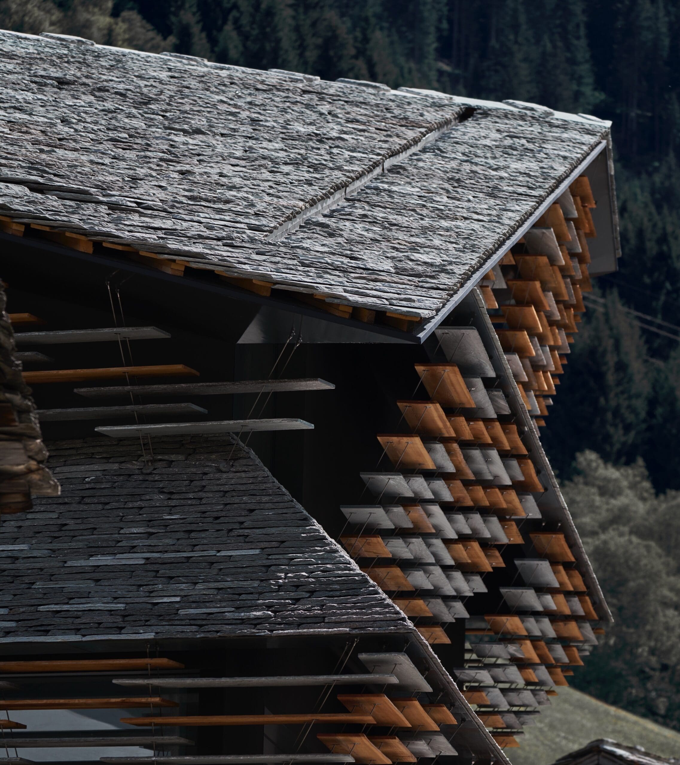 Close-up of modern buildings with overlapping Vals Quartzite stone shingle roofs and wooden slats, set against a blurred forested mountain background.