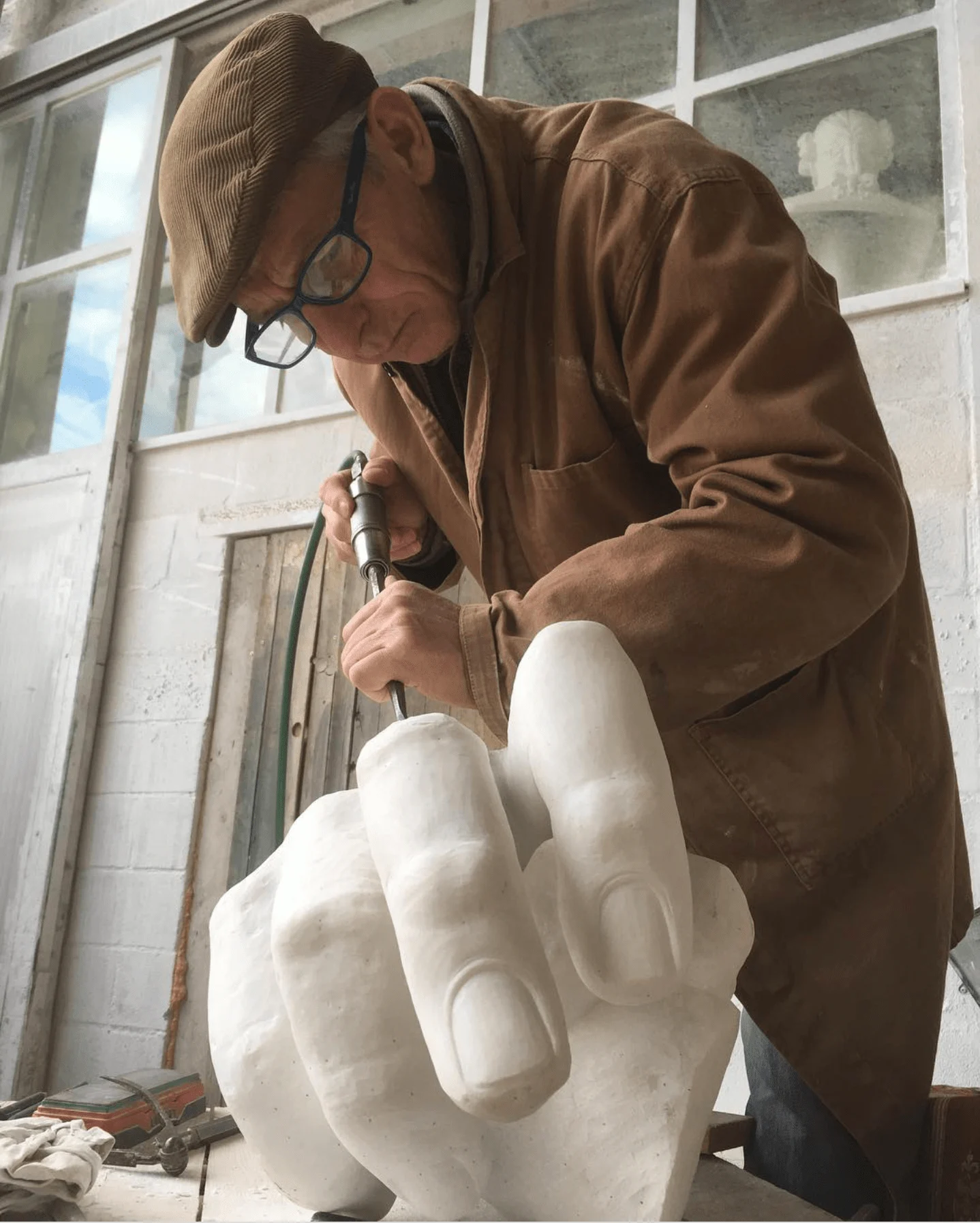 Elderly man wearing a flat cap and glasses chiseling a large white stone sculpture resembling a hand. Hes focused on his work, with various tools visible around him. The space appears to be a workshop with white walls and a window.