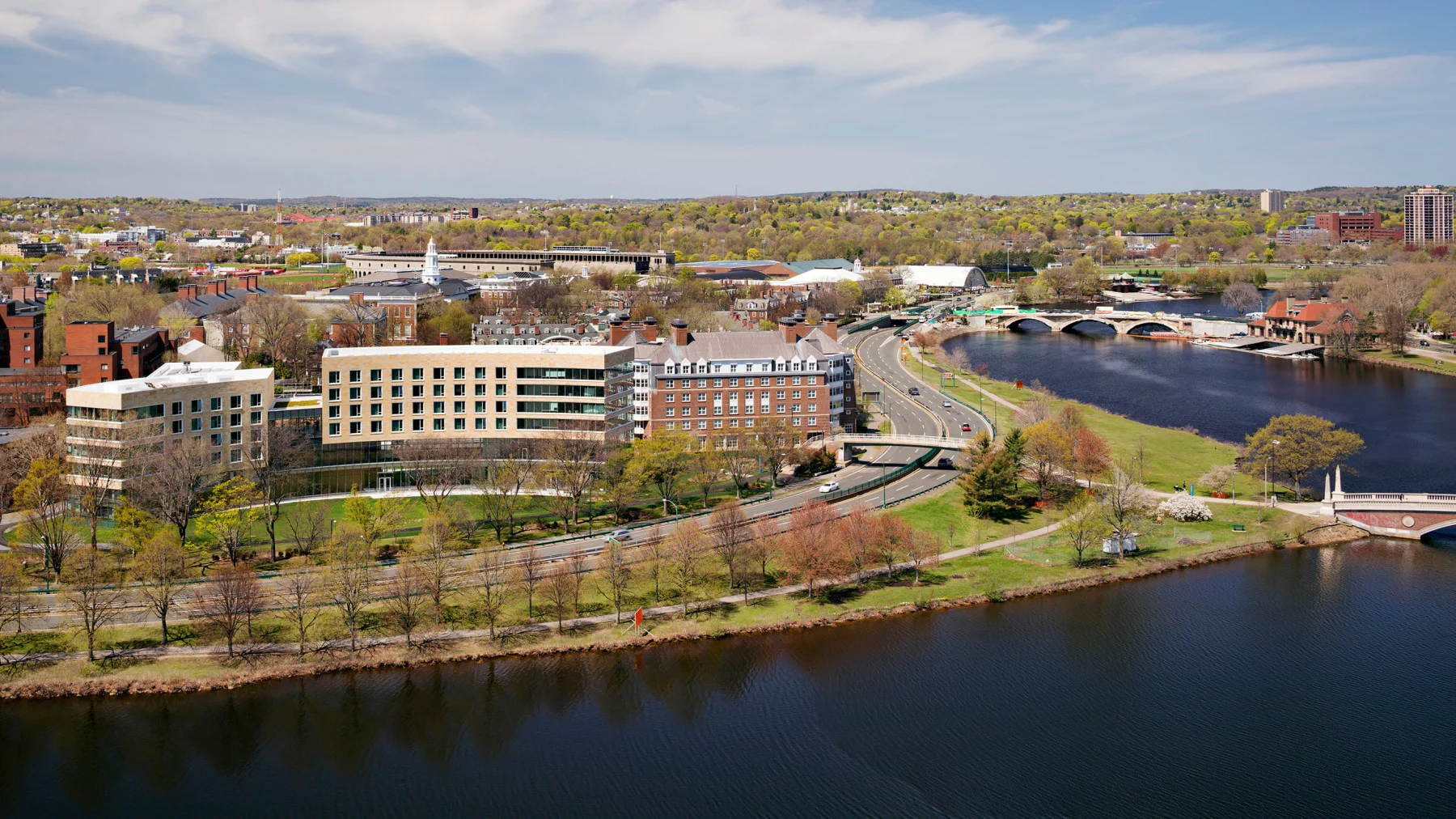 Aerial view of a university campus by a river, featuring modern and traditional buildings, a bridge, and a tree-lined walkway. The sky is partly cloudy, and the surrounding landscape includes greenery and distant structures.