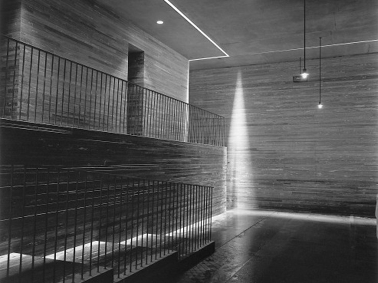 A minimalist interior with concrete walls and metal railings features dramatic lighting that casts geometric shadows. Light streams in, accentuating the natural stone texture of the Vals Quartzite and highlighting the modern architectural design.