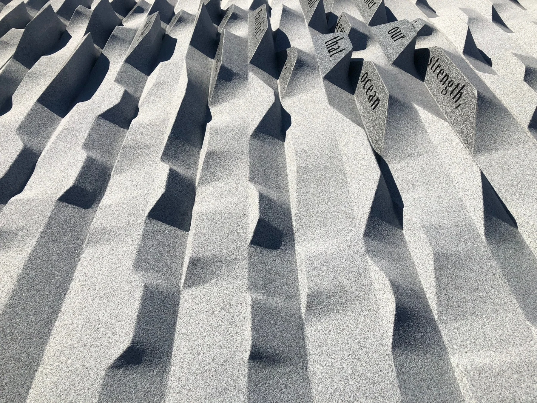 A textured sculpture featuring multiple wavy, vertical patterns. The surface is gray, with shadows accentuating the ridges. Words such as strength and ocean are partially visible on some of the raised areas.