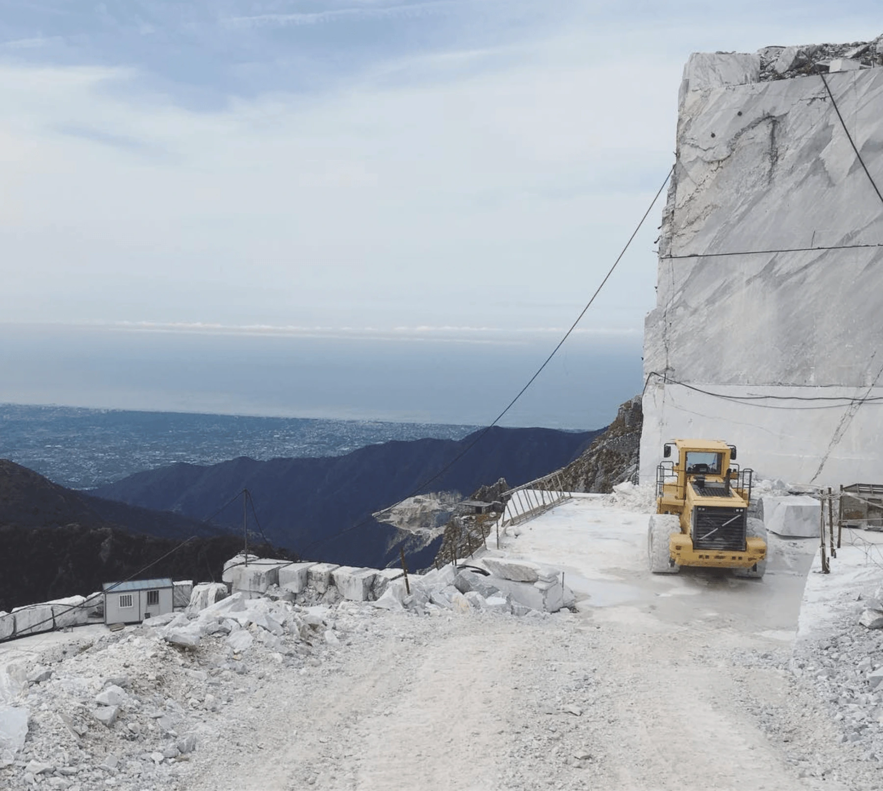 A yellow bulldozer works in a large marble quarry, surrounded by rugged mountains. The quarrys white stone contrasts with the distant view of a city and the sea under a cloudy sky.