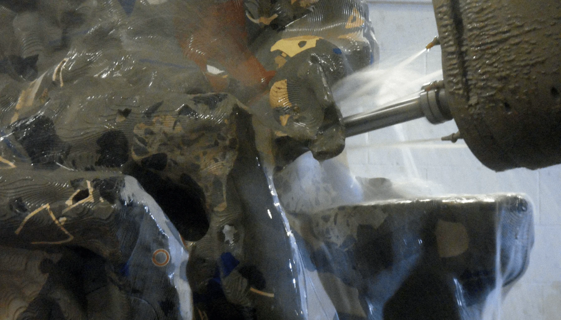 A CNC machine cuts into a large, abstract sculpture made of different materials. The sculpture has a shiny, multi-colored surface. Water or coolant is being sprayed onto the area where the machine is working.