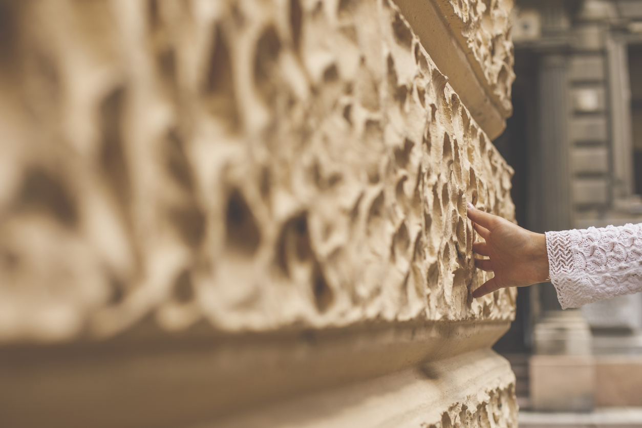 A person wearing a white lace sleeve touches a textured, beige stone wall with their fingertips, highlighting the transformation made possible by Prestige Stone Restoration. The background reveals soft-focused architectural details.
