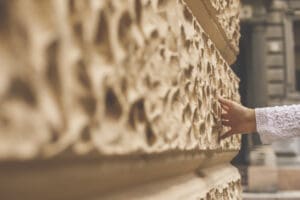 A person wearing a white, lacy sleeve touches the textured, beige stone wall of a historic building, with architectural details visible in the blurred background.
