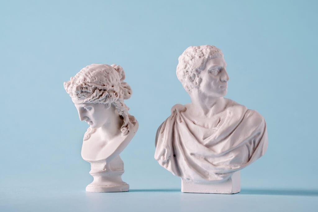 Two white marble sculptures—a classical bust of a woman with braided hair and a man in a toga—are displayed side by side against a light blue background, blending the elegance of classical art with modern simplicity.